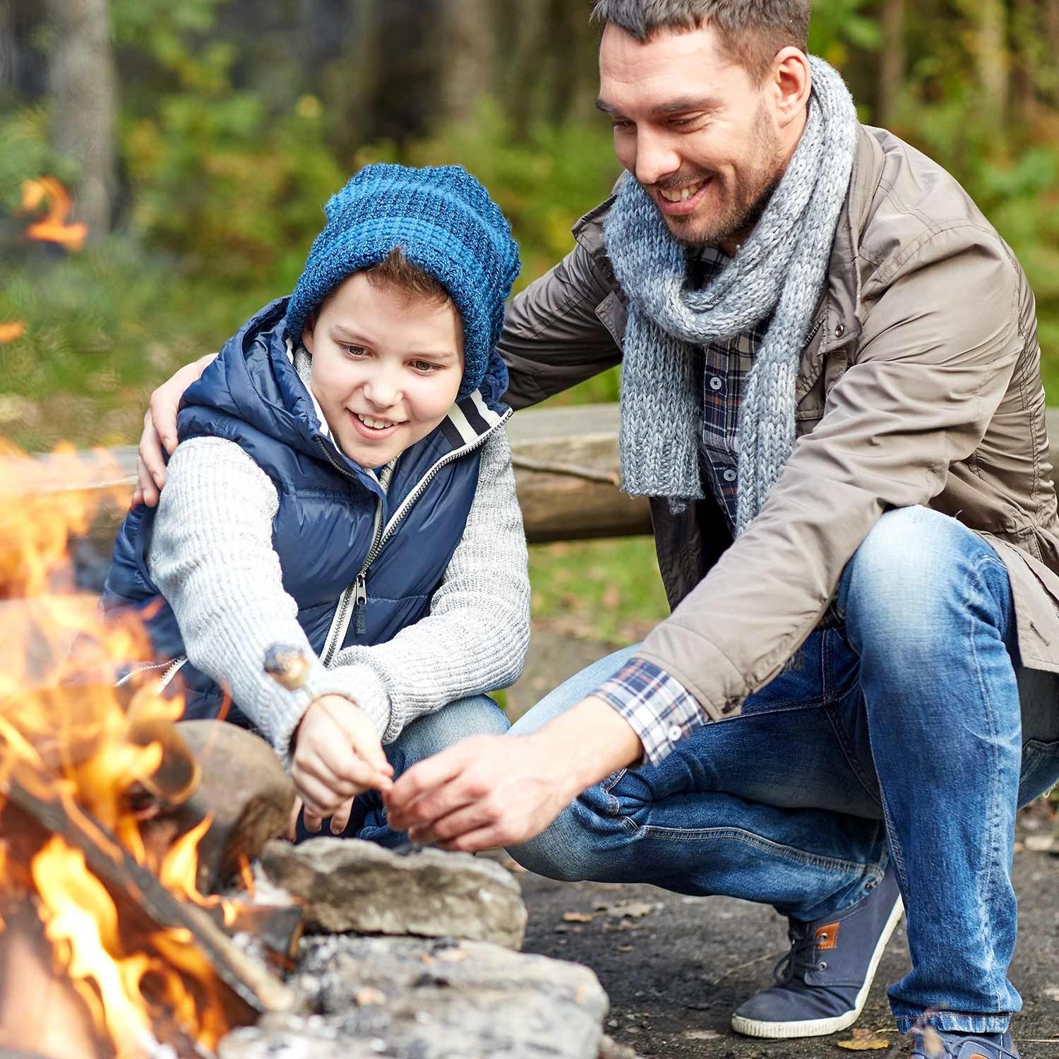 A man and a child are roasting marshmallows over a campfire in an outdoor setting at Montebello VA camping, both smiling and wearing warm clothes.