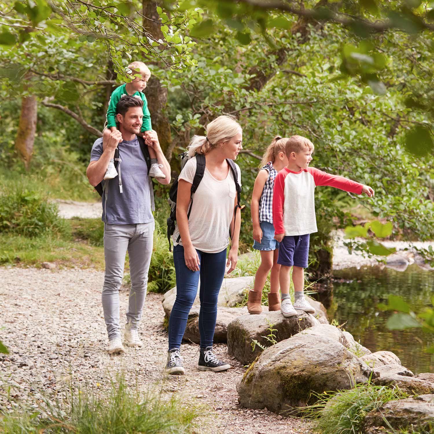 A family of four, with two kids, walks along a forest path near a pond during their Montebello VA camping trip. One child points at the water, while the other sits on an adult’s shoulders. Everyone looks relaxed and happy, enjoying nature.